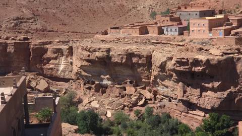 Steep rocky canyon walls with small cliff dwellings and a village perched above a green riverbed.