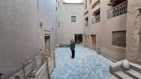 Woman stands in a narrow stone-paved kasbah alley flanked by high adobe walls and wooden balconies.