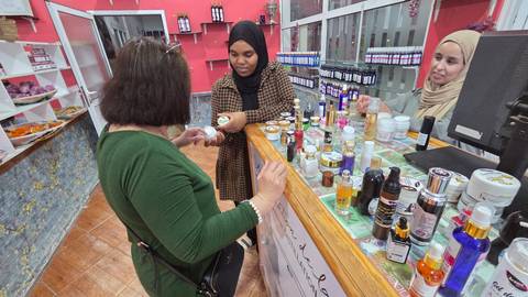 Tourist samples cosmetics from saleswomen behind a colorful counter in a small shop.