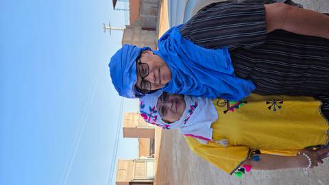 Couple dressed in traditional scarves smile together outside desert buildings under clear blue sky.