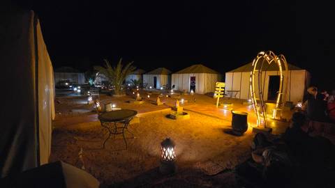 Night scene of luxury desert camp with canvas tents, palm tree and dozens of lanterns lighting sandy paths.