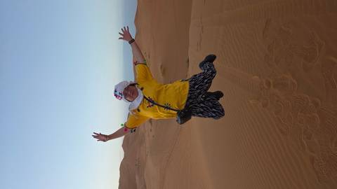 Traveler captured mid-air jumping joyfully on soft orange dunes against pale evening sky.