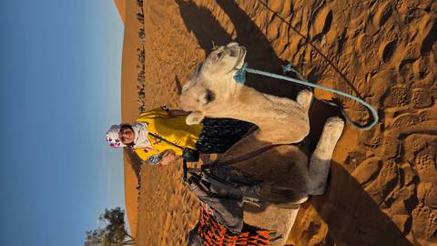 Traveller standing beside saddled camel on sun-lit sand dunes under clear blue sky.