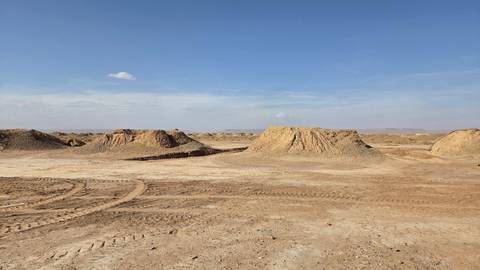 Barren desert landscape with low earthen mounds and tyre tracks under clear sky.