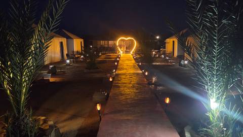 Nighttime desert camp walkway lit by lanterns leading to heart-shaped light arch among tents.
