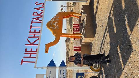 Visitor standing beside bright camel statue under sign 'The Khettaras' in desert area.