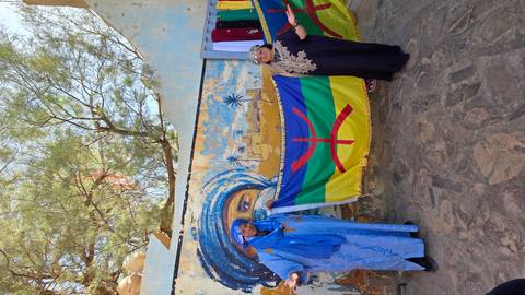 Two travellers in traditional dress holding Amazigh flag before a painted desert mural.