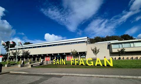 Modern entrance building of St Fagans National Museum with bold yellow signage under a vivid blue sky.