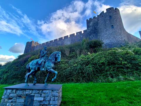 Bronze equestrian statue stands before the stone battlements of a medieval Welsh castle against a bright sky.