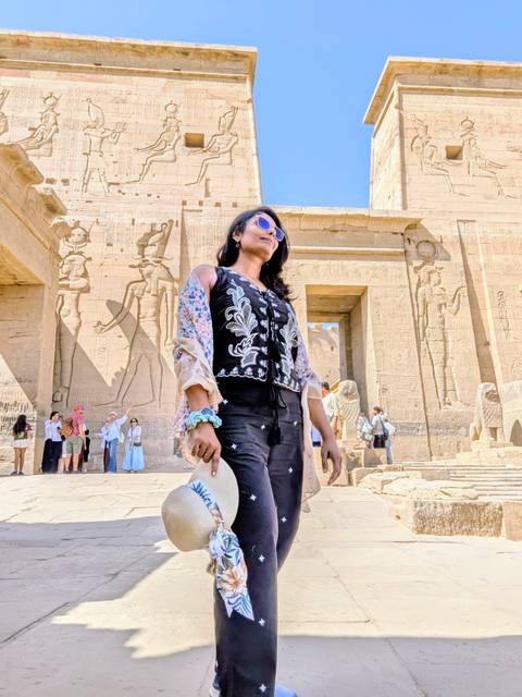 A woman wearing sunglasses poses in front of a sandstone Egyptian temple wall covered with large ancient relief carvings while other visitors mill about in the background.