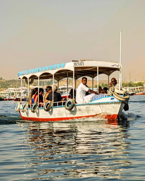 A small covered motorboat full of tourists cruises along a wide river with desert hills in the hazy distance.