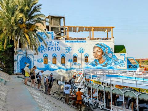 Tourists disembark beside a brightly painted Nubian riverside building featuring a blue mural of a smiling woman and Egyptian motifs.