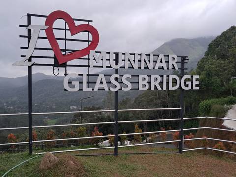 Large sign reading 'I ♥ Munnar Glass Bridge' set against misty green mountains