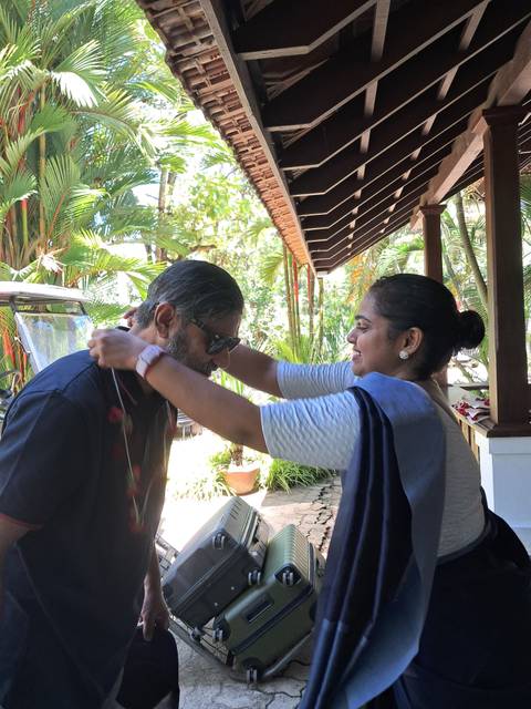 Woman placing a welcome garland on a guest at a tropical resort entrance surrounded by palms