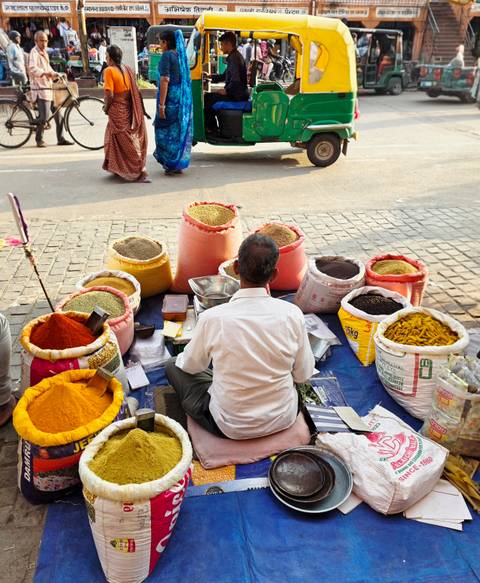 Street vendor seated on the ground surrounded by colourful open sacks of spices and grains