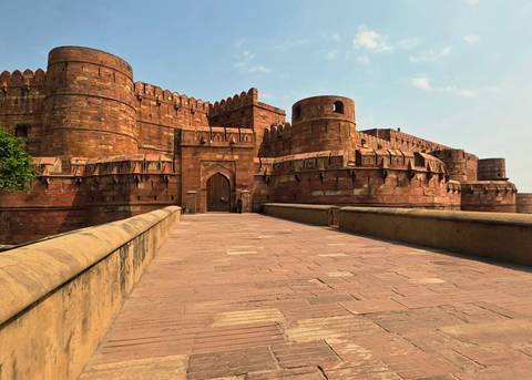 Red‐sandstone fort walls with arched gateway viewed from an empty stone causeway under a clear blue sky.