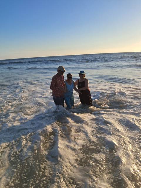 Three adults laughing while standing in gentle surf at sunset beach.
