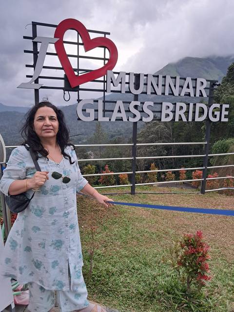 Woman holding sunglasses beside a sign reading "Munnar Glass Bridge" against misty hills.