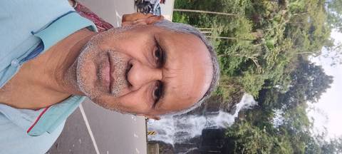 Selfie of a traveller with a roadside waterfall cascading behind him in a lush forested area.