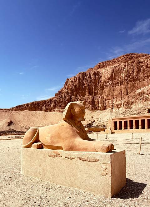 Stone sphinx statue with dramatic red cliffs and an ancient temple facade in the background.