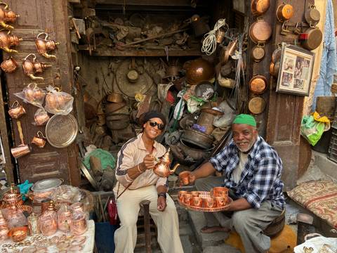 Traveller sharing tea with a copper artisan inside a bustling Moroccan souk stall.