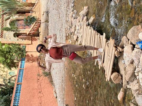 Traveller flexing on a makeshift wooden footbridge over a shallow mountain stream near an adobe village.