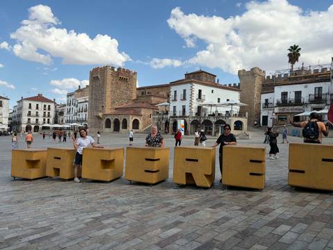 Tourists posing with large yellow block letters spelling out a city name in a historic Spanish plaza with medieval towers