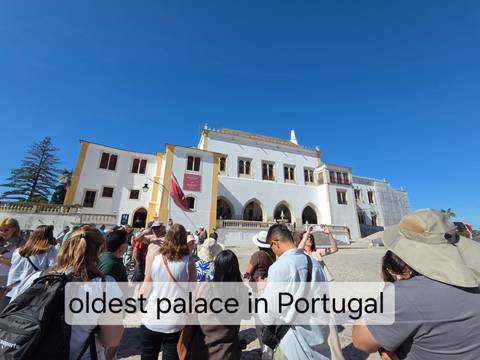 Large tour group gathered outside a historic white and yellow Portuguese palace under clear blue sky with caption bar