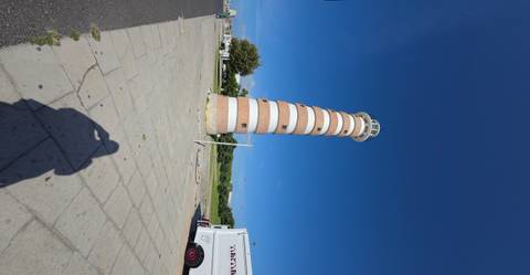Tall striped brick lighthouse rising against a deep blue sky, photographed from ground level with long shadow in foreground