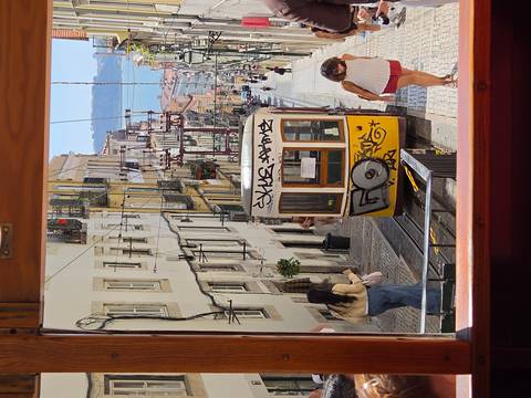View through a tram window of Lisbon’s steep Elevador da Bica track with yellow funicular, pedestrians and distant river