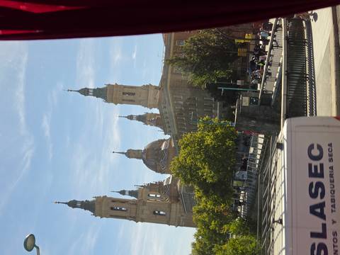 View from a moving vehicle of the Basilica of Our Lady of the Pillar’s towers rising behind trees and urban buildings