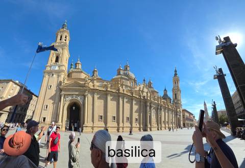 Wide-angle view of Basilica del Pilar with many tourists and partial text overlay at the bottom edge
