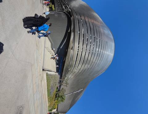Modern metallic museum building with sweeping curves, groups of visitors on broad steps beneath clear sky