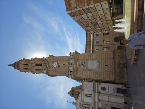 Stone clock tower back-lit by the sun with a fountain in the plaza and a smiling traveler in foreground
