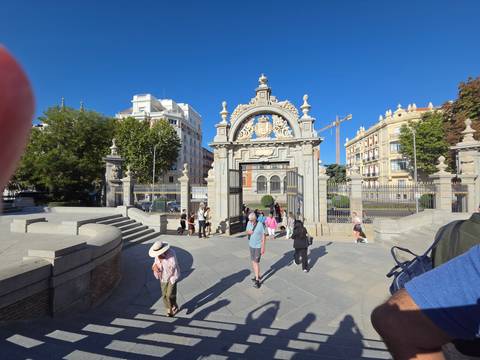 Decorative wrought-iron gate entrance to a city park with tourists milling around; a finger intrudes on upper left