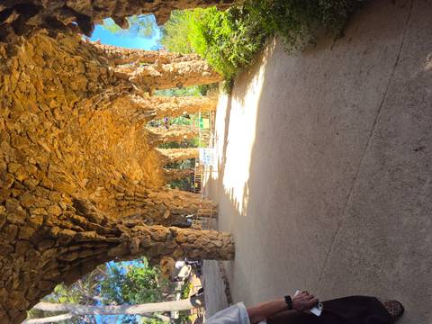 Rustic stone arch structures in Park Güell with bright sunlight filtering between pillars and safety barriers present