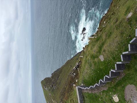 Steep grassy cliff dropping to the Atlantic with waves crashing below under gray skies