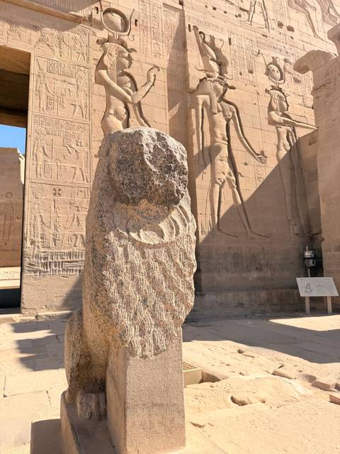 Weathered granite statue fragment stands before hieroglyph-covered sandstone wall.