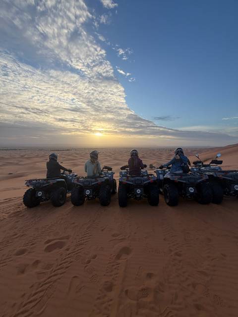Five travellers on quad bikes look over sweeping Sahara dunes at sunset.