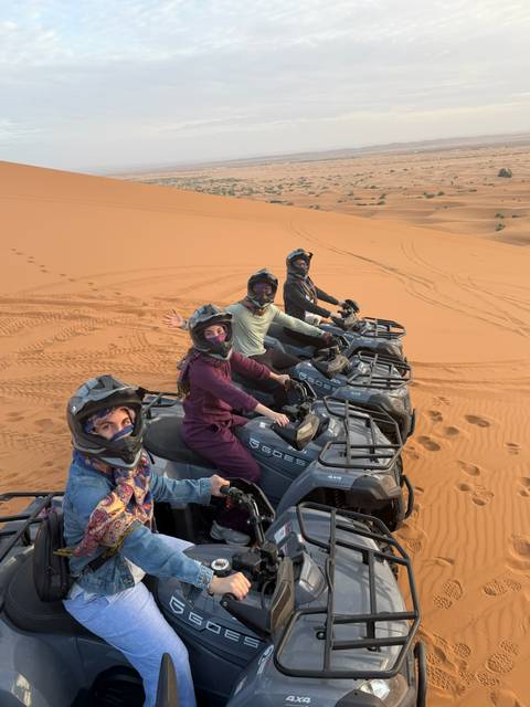 Riders grin from their quad bikes lined up on patterned desert sand.