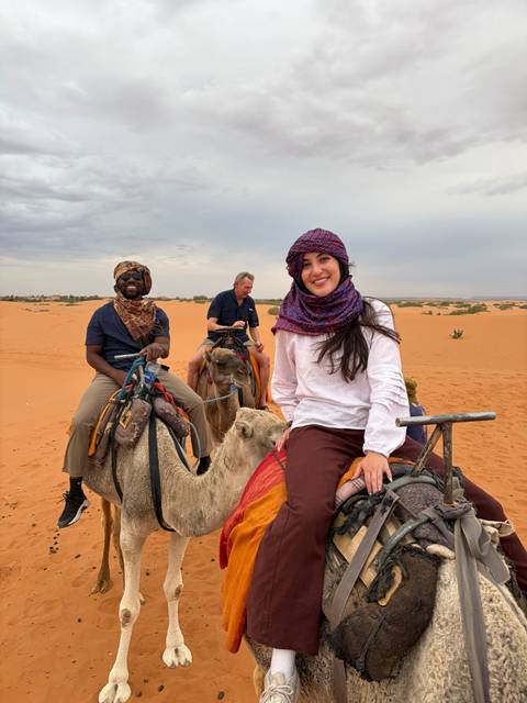 Travellers ride camels through orange desert sands, smiling beneath colorful scarves.