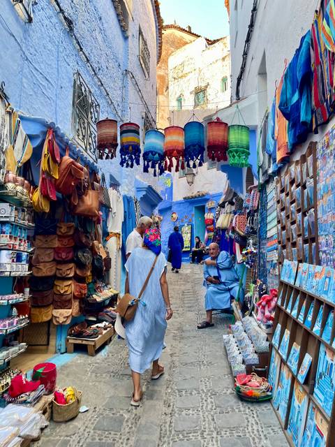 Colorful market lane in the blue-washed medina lined with leather bags and textiles, shoppers and vendors in traditional dress.