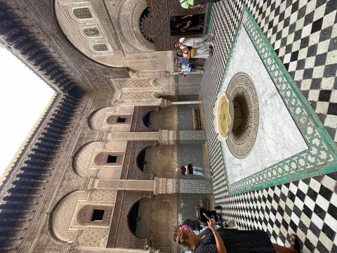 Mosaic-tiled courtyard with a marble fountain in a historic medersa, visitors standing along the walls.
