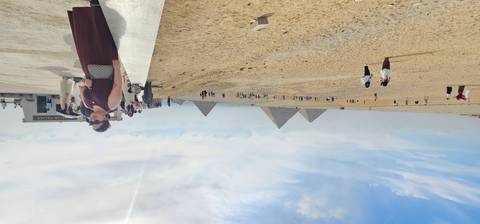 Wide desert panorama with distant pyramids and scattered tourists under hazy sky.