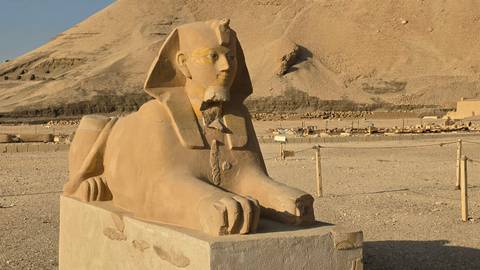 Stone sphinx statue sits before desert hills and ancient ruins in warm sunlight.