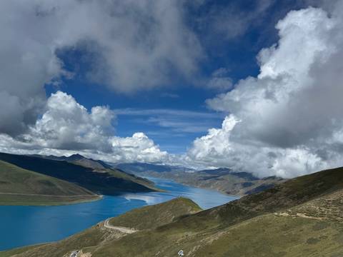 Deep-blue Yamdrok Lake winds between green and brown Tibetan mountains beneath billowing clouds.