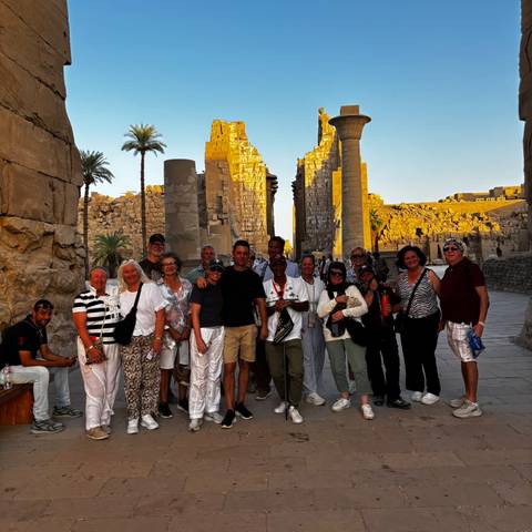 Large tour group stands among sunlit columns and obelisks of an ancient Egyptian temple at sunset.