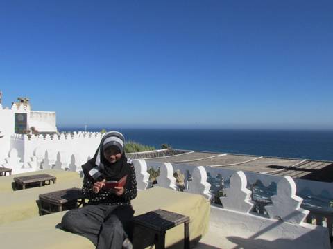 Young woman in hijab reads on a rooftop terrace overlooking a vast blue sea.