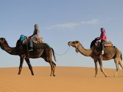 Two women ride camels across orange Sahara sand dunes under blue sky.