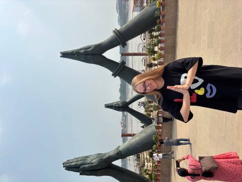 Traveler making a namaste gesture in front of giant praying-hand sculptures beside a river